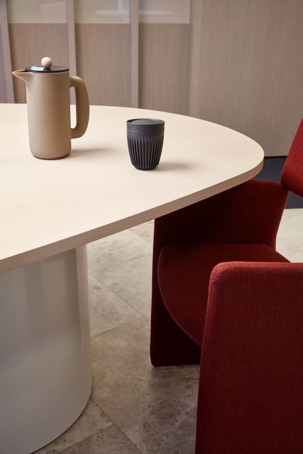 Close-up interior view of a refined dining area featuring a pale oval table with a rounded pedestal base, two deep red upholstered dining chairs, a matte beige carafe, and a dark ribbed cup. The room has warm light-wood wall panelling with pale trim and large beige stone-look floor tiles.