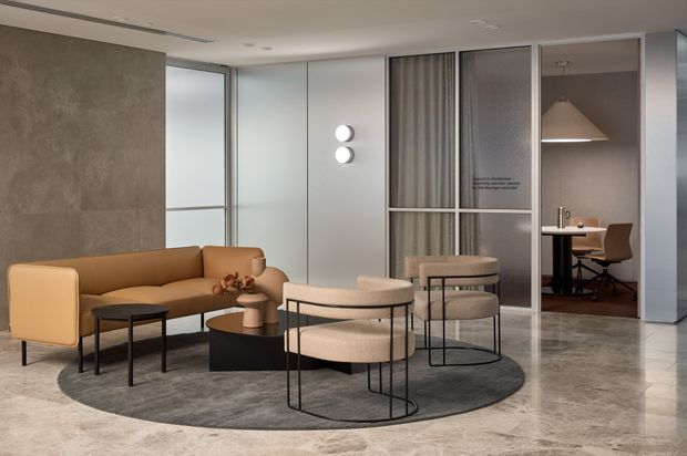 A refined contemporary lounge and reception area with a tan upholstered sofa, rounded boucle-style lounge chairs, small dark coffee tables, and a large circular grey rug. Full-height glazed and framed partitions create a visible home office or meeting room beyond, with a desk and task chair. The space features polished stone flooring, neutral grey wall finishes, concealed linear ceiling detailing, and soft architectural lighting.