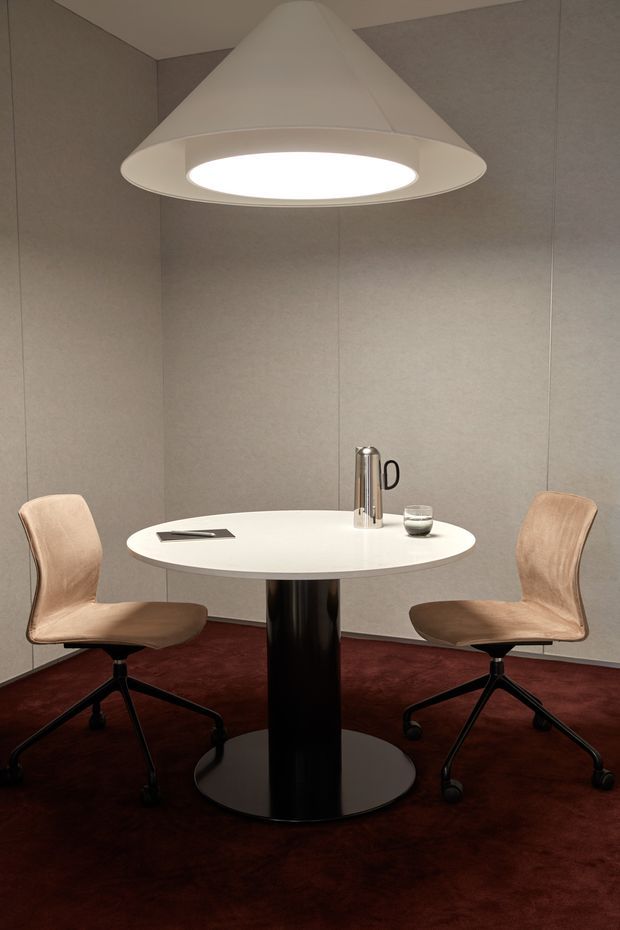 A compact, enclosed meeting or dining nook with a round white table on a black pedestal, two upholstered swivel office chairs, and a large white cone-shaped pendant light. Neutral grey wall panels surround the space, while a deep burgundy carpet adds contrast. Minimal tabletop accessories and dark furniture details create a refined, intimate atmosphere.