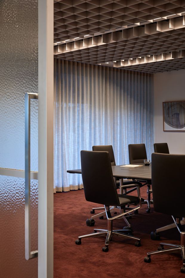 A refined contemporary conference room viewed through a partially open glass door. The room contains a long dark meeting table surrounded by black leather swivel chairs, with full-height sheer curtains, a deep burgundy carpet, textured glass and a sculptural gridded ceiling with integrated pendant-style lighting. The palette is warm, dark and sophisticated, with strong geometric architectural detailing.