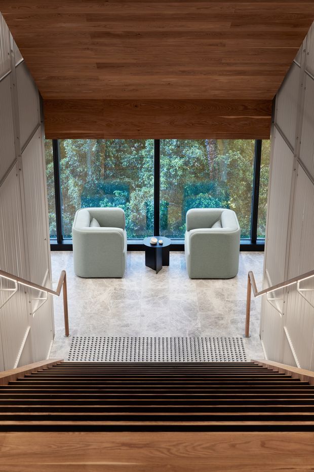 A refined interior stairway viewed from above, descending toward a small seating area framed by expansive black-framed glazing. The stair and ceiling are finished in warm timber, while the side walls feature pale vertical ribbed panelling with integrated linear lighting and timber handrails. At the landing, two light upholstered armchairs and a compact dark side table sit on a pale stone floor, with leafy vegetation visible beyond the windows.