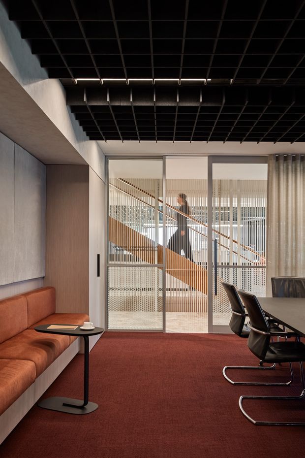 A refined contemporary commercial interior, likely a meeting or lounge area, with a rust-red upholstered banquette, dark meeting table and cantilevered chairs, deep red carpet, timber wall panelling, and a dramatic black open-grid ceiling. Full-height glazed doors reveal a sculptural timber staircase and vertical screening beyond, while sheer curtains soften the right side of the room.