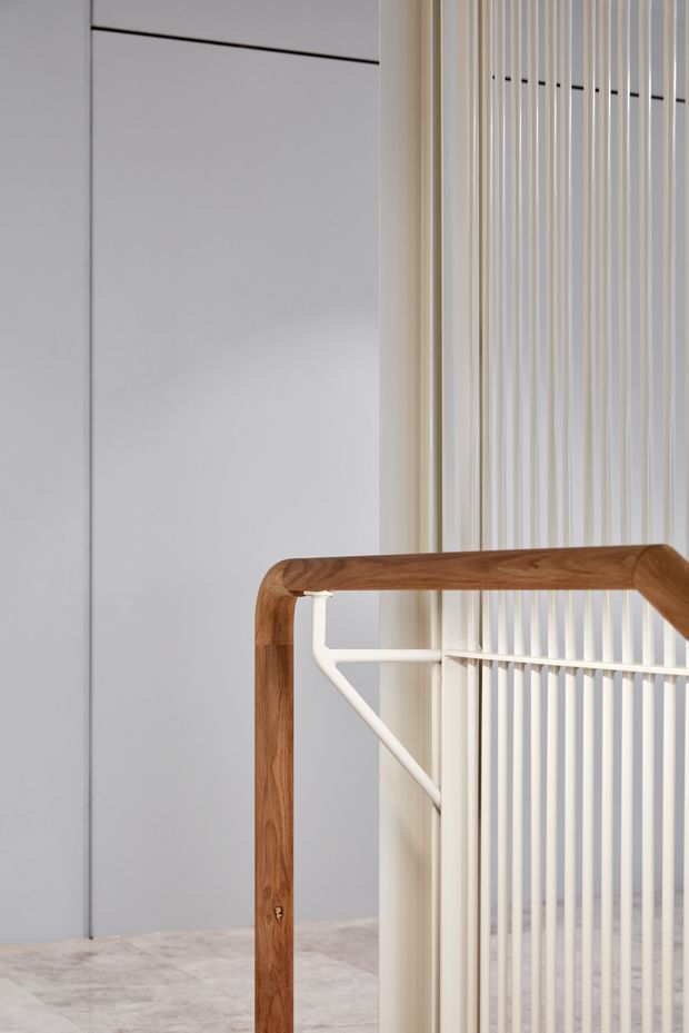 Close-up architectural detail of a minimalist stair or landing with a warm timber handrail, white metal support bracket, vertical white balustrade slats, pale built-in wall panels, and light stone or concrete flooring. The composition emphasizes clean lines, contrasting timber and white finishes, and refined contemporary detailing.