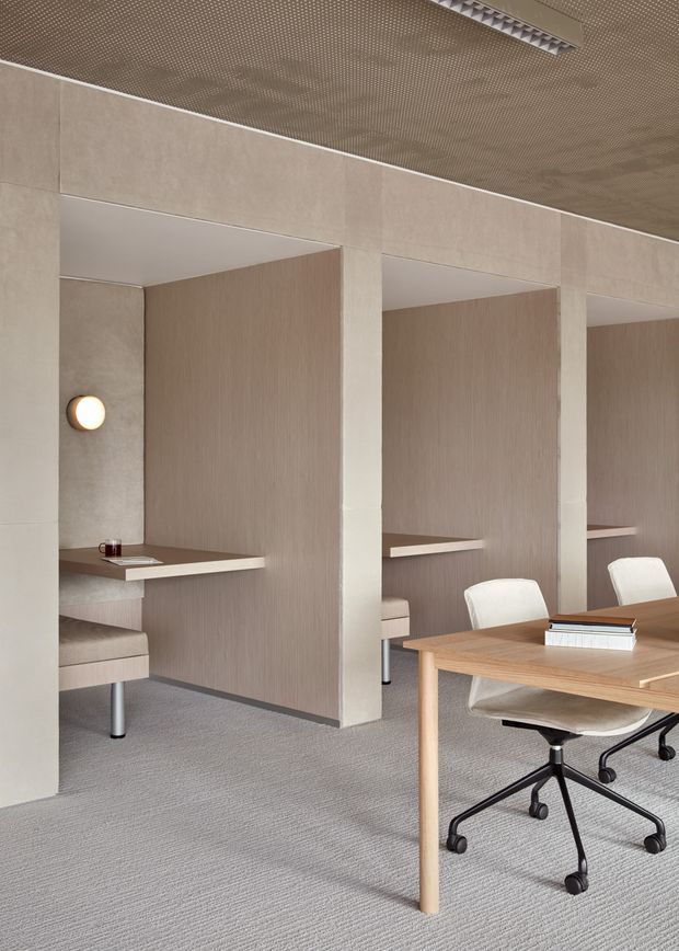 A refined contemporary office interior with multiple semi-enclosed individual work booths, pale textured wall panels, integrated desks, upholstered swivel task chairs, and a shared timber table in the foreground. The space has a muted neutral palette, grey carpet, warm wood tones, and a perforated acoustic ceiling with linear lighting.