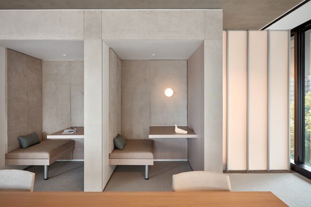 A refined contemporary interior featuring a built-in study or work nook within a concrete-toned alcove. The space includes a fixed desk, upholstered bench seating, a small wall light, full-height panelled walls, and a large mirror reflecting the adjacent nook. Vertical translucent screens and expansive black-framed glazing bring soft daylight into the room. A timber table occupies the foreground.