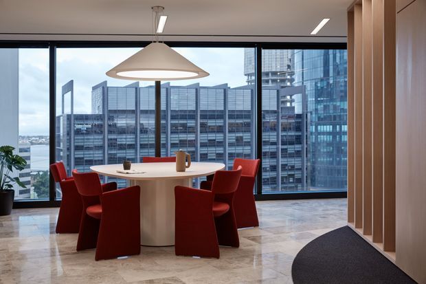 A refined high-rise dining or breakout area with a round white table surrounded by vivid red upholstered chairs. Floor-to-ceiling windows provide expansive views of a dense urban skyline and water beyond. The space features polished light stone flooring, warm timber vertical wall panelling and joinery, black-framed glazing, linear recessed lighting, and a large sculptural pendant above the table. A dark entrance mat and potted plant complete the scene.