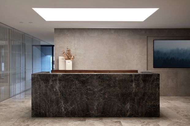 A refined contemporary reception or entry space dominated by a large dark veined stone-clad counter or island. Behind it is a warm grey textured feature wall with a recessed wall-mounted screen, timber ledge, and simple ceramic vessels. Full-height fluted or ribbed glass partitions line the left side, while a large geometric recessed ceiling light provides even illumination. The floor is finished in pale stone or porcelain tile.
