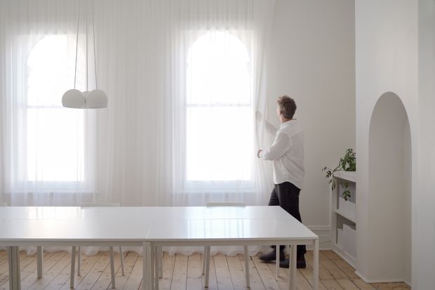 Bright, airy dining area with a long white dining table, pale timber plank flooring, sheer floor-to-ceiling curtains, tall arched windows, a sculptural white pendant light, and a white arched built-in shelving niche. A person is standing by the window, and a small plant adds greenery.