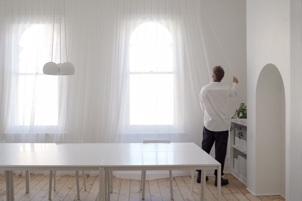 Bright, airy white interior with tall arched windows dressed in sheer curtains, pale timber flooring, a long white dining or work table, and a person adjusting a curtain beside an arched wall opening. Minimal built-in shelving and a small plant add subtle detail.