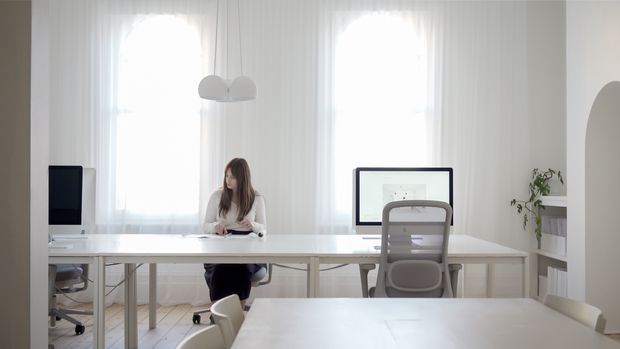 Bright, airy contemporary office or studio workspace with long white desks arranged in rows, a woman working at a workstation, ergonomic office chairs, computer monitors, sheer floor-to-ceiling curtains, pendant globe lighting, and open white shelving with a small plant. The predominantly white interior has abundant natural light and a clean, minimal fit-out.