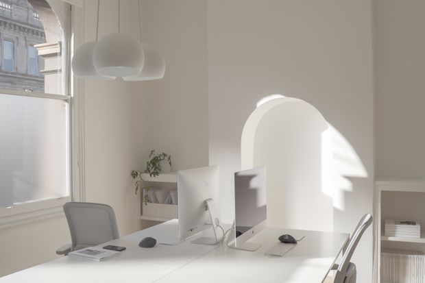 Bright minimalist office interior with two white computer workstations arranged on a shared desk, ergonomic mesh chairs, a sculptural white pendant light, open shelving, and tall windows. Strong sunlight creates an arched geometric shadow on the white wall, while an ornate neighbouring building is visible outside.