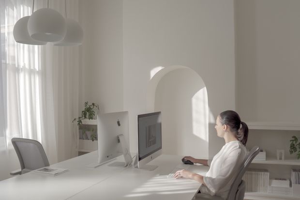 Bright minimalist home office with a long white desk, dual computer workstations, ergonomic chairs, sheer floor-to-ceiling curtains, sculptural white pendant lights, and a rounded arched opening. Soft daylight creates a calm, refined atmosphere, with sparse shelving and indoor plants adding subtle warmth.