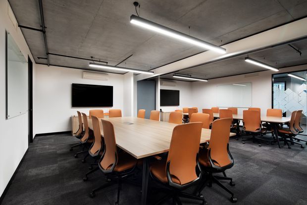A modern commercial conference and meeting room with a large light-wood boardroom table surrounded by orange ergonomic office chairs. The space features dark carpet flooring, white walls, exposed concrete ceilings, black linear pendant lighting, wall-mounted screens, and glazed partitions.