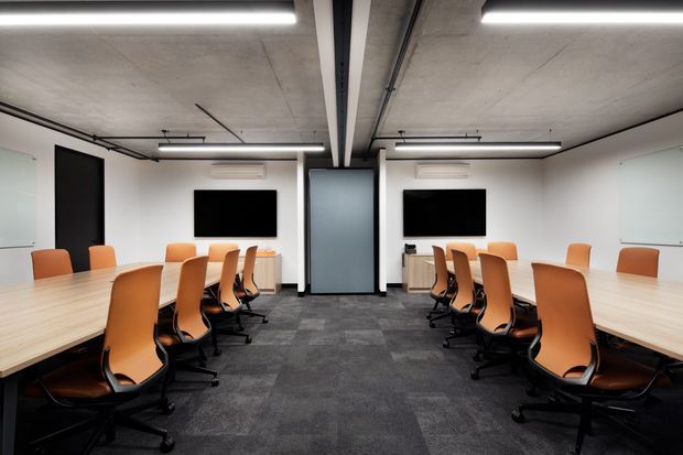 A contemporary commercial boardroom or meeting space with two long light-wood conference tables arranged on either side of a central glazed partition. Orange ergonomic office chairs surround the tables, with wall-mounted black displays, white walls, exposed concrete ceilings, suspended linear lighting, and dark grey carpet flooring.