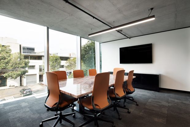 Modern meeting room with a long polished stone conference table surrounded by orange-brown ergonomic office chairs. Floor-to-ceiling glazing provides abundant daylight and views of an urban streetscape. The room has a raw exposed concrete ceiling, linear suspended lighting, dark carpet tiles, white walls, and a wall-mounted television above a dark credenza.