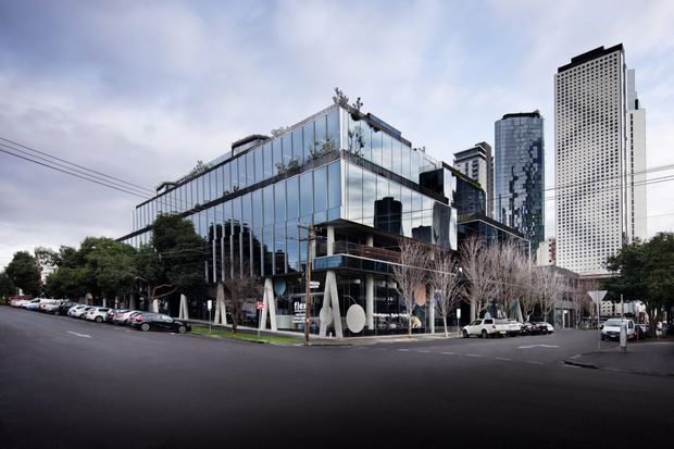 Street-level view of a contemporary urban commercial building with extensive reflective glass curtain walls, landscaped terraces, and distinctive angled structural columns. Tall office towers rise behind it, with a broad paved street and parked vehicles in the foreground.