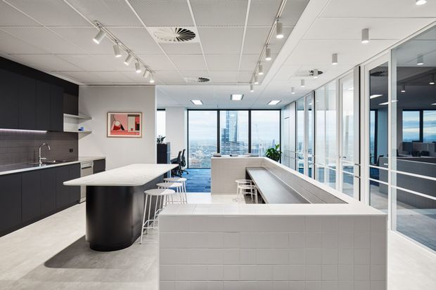 A contemporary commercial office breakout and kitchenette area with a large black island, dark integrated cabinetry, tiled splashback, and white stone-look surfaces. A long tiled breakfast bar with stools separates the kitchen from an open collaborative workspace. Full-height glazed partitions enclose meeting rooms, while city views are visible through the windows. The ceiling has recessed services and track lighting, creating a bright, professional workplace interior.