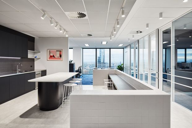A bright contemporary commercial office breakout and kitchenette area with a long tiled island or counter, bar stools, dark integrated cabinetry, and a large communal island. Full-height glazed partitions reveal meeting rooms and a city skyline beyond. The space has a clean, open-plan layout with linear track lighting and neutral finishes.