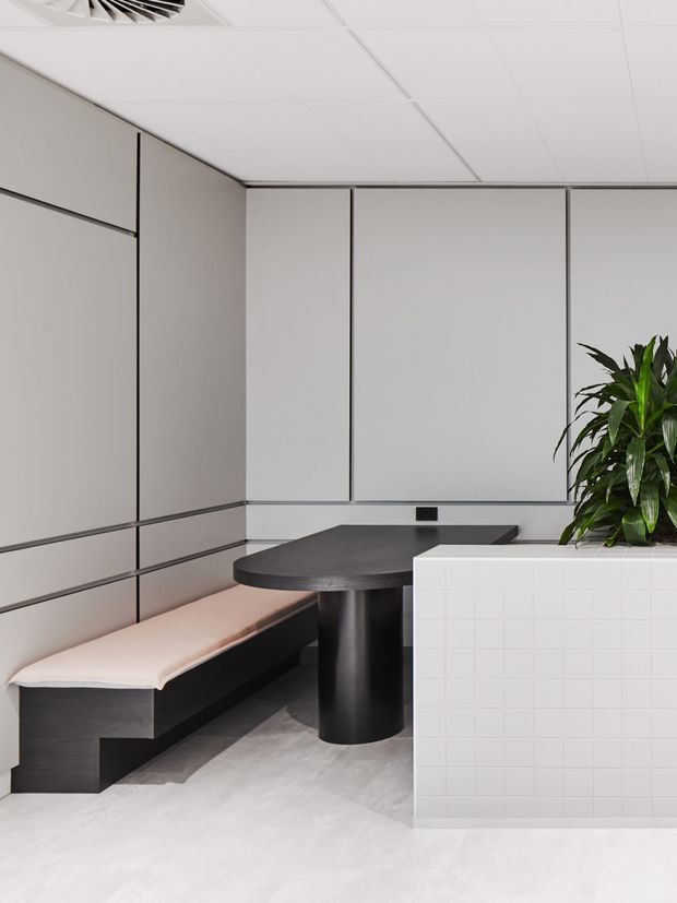 A compact, refined commercial or office-style breakout area with a built-in upholstered bench wrapping around a dark sculptural table. Full-height light grey wall panels with slim dark reveals create a minimalist backdrop, while a white tiled planter or partition with a leafy indoor plant defines the foreground. The space has a suspended white acoustic ceiling and pale flooring.