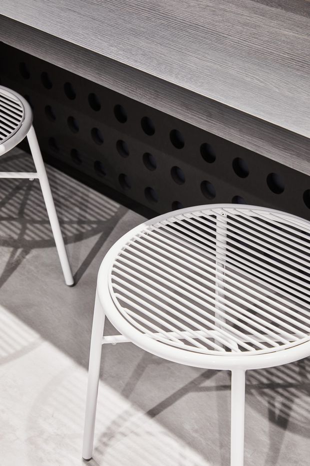 Close-up of two white round slatted outdoor-style stools or side tables positioned on a light grey floor beside a dark perforated wall panel and a horizontal grey wood-grain surface. Strong sunlight creates striped shadows from the slatted furniture. The image is a detail-focused product or furniture composition with a clean contemporary look.
