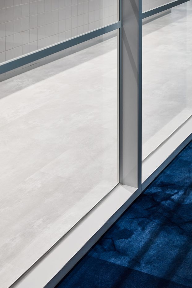 Close-up architectural detail of a glazed partition or doorway with slim dark metal framing, pale large-format floor tiles, white tiled wall, and a deep blue carpeted area beyond the threshold. The image emphasizes the junction between hard flooring, glazing, and carpet.
