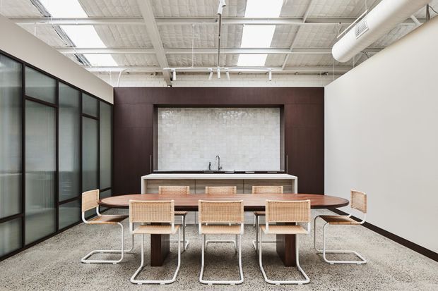 A contemporary open-plan dining and kitchen area with a long wooden dining table, six woven-seat cantilever chairs, and a compact kitchen bench set within dark timber cabinetry. A pale textured stone backsplash, black-framed glazed partition, polished concrete floor, and exposed white services create an industrial office or hospitality-style interior.