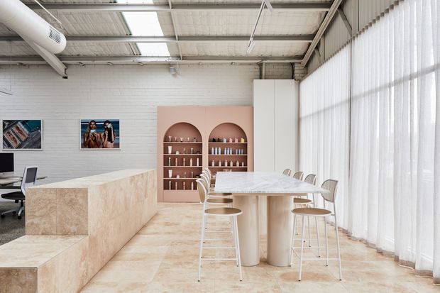 Bright open-plan commercial interior with an industrial warehouse shell, white painted brick walls, exposed metal roof structure, tall sheer-curtained glazing, and pale stone-look finishes. A large island counter and integrated bench seating sit beside a communal dining table with stools. Arched built-in shelving displays bottles and products, while a small workstation is visible at left.
