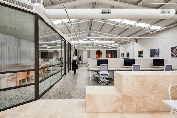 A bright contemporary open-plan office with exposed white structural roof framing, ductwork and services, extensive black-framed fluted-glass partitions, rows of desks with computer monitors, ergonomic office chairs, and a large pale stone reception or workstation feature. Polished concrete or terrazzo-like flooring and artwork add visual interest.