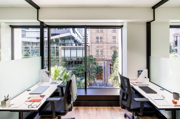 Bright, contemporary shared office workspace with two facing white desks, ergonomic black office chairs, computers and stationery. Full-height windows provide abundant natural light and views of surrounding urban buildings, while black-framed glazing and pale timber flooring define the clean, professional interior.