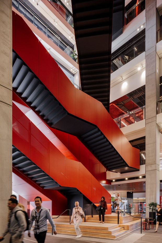 Interior view of a large contemporary multi-level commercial atrium dominated by a dramatic zigzagging staircase. The stairs feature bold red perforated and solid balustrade panels with dark metal treads and undersides, connecting several floors around a tall central void. Exposed concrete columns, glass balustrades, linear lighting, timber-toned stepped seating, and people moving through the lobby are visible.