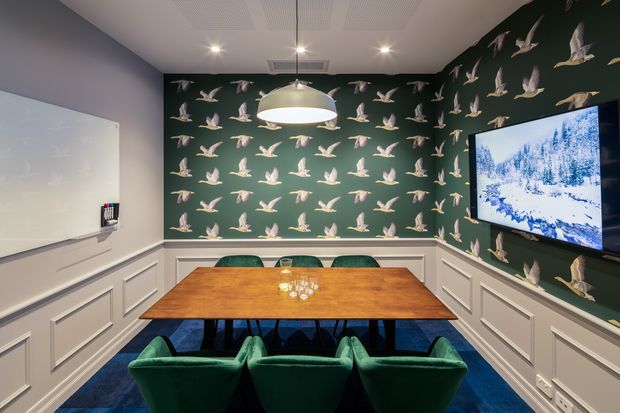 A compact, richly decorated dining room or meeting space with a large rectangular timber table, green upholstered chairs, dark green bird-pattern wallpaper, white decorative wall panelling, a wall-mounted television, and a large white pendant light. A whiteboard is mounted on the left wall, suggesting the room may also function as a boardroom or collaborative workspace.
