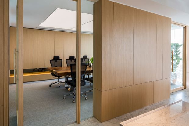 A refined contemporary office meeting room viewed through a full-height glazed partition and timber-framed door. The room has extensive light timber wall panelling and built-in cabinetry, a large rectangular timber meeting table with black office chairs, grey carpet, and a recessed linear bench with warm lighting. A large square skylight or ceiling light brightens the room, while a marble-lined corridor and a planted area are visible outside the meeting room.
