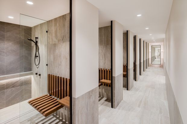 A bright, contemporary bathroom and corridor viewed through a series of open shower-room bays. The foreground shows a walk-in shower with a black rain shower, large grey-beige stone-look wall tiles, a timber slatted folding seat, and a dark vertical radiator. Repeated shower cubicles line the long corridor, creating strong perspective and rhythm, with recessed ceiling lights and pale stone-look flooring throughout.
