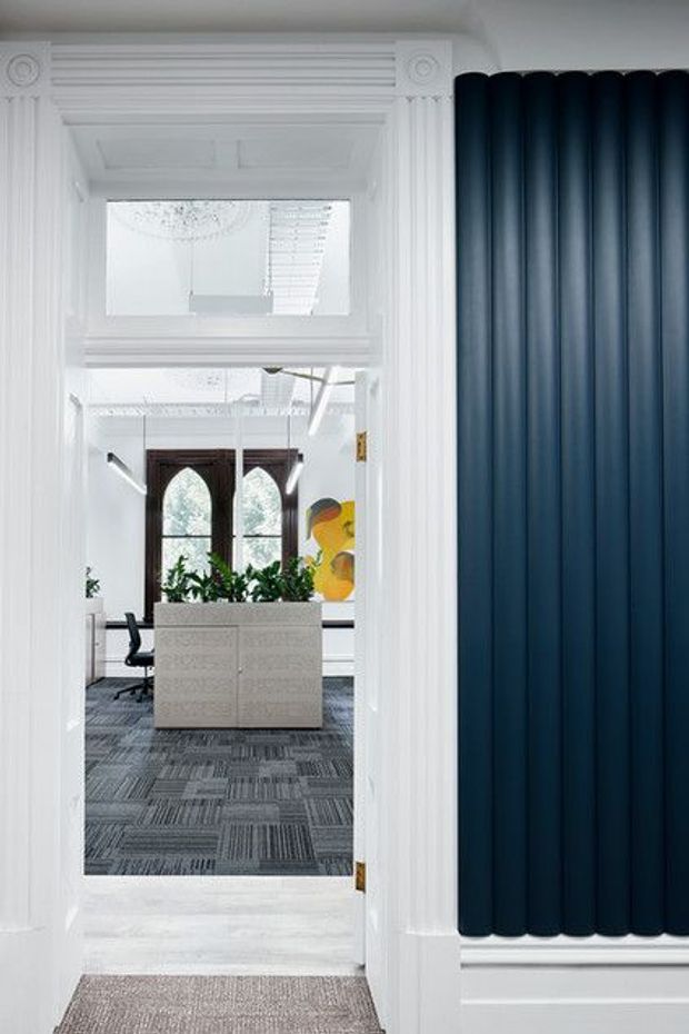 View through an ornate white-trimmed doorway into a bright, refined office or work area. The foreground wall features a dramatic dark navy fluted panel, while the adjoining room has patterned carpet tiles, a pale central planter or storage unit with greenery, office seating, tall arched timber doors, contemporary linear lighting, and a colourful artwork. Decorative classical mouldings and ceiling details contrast with modern furnishings.