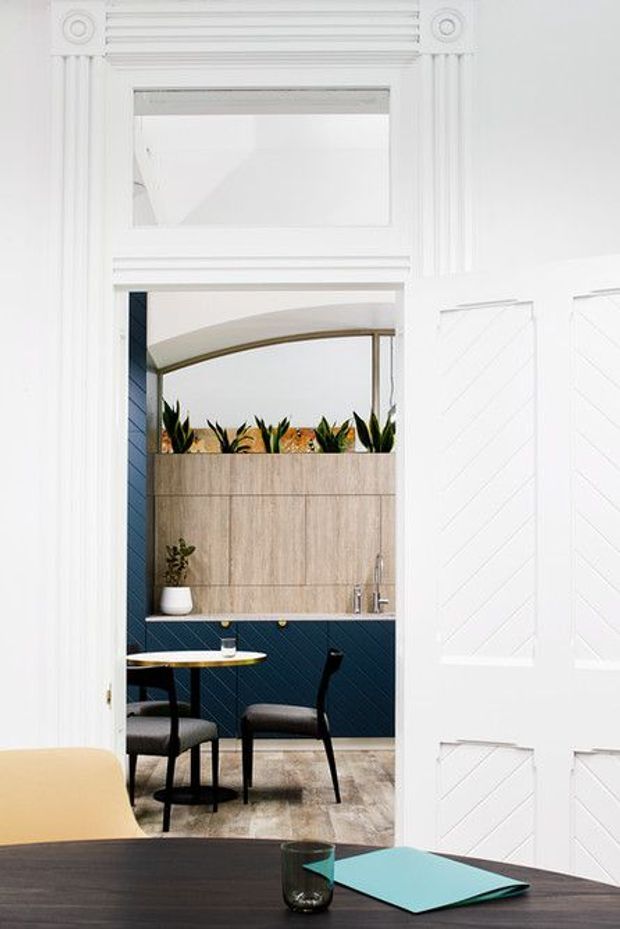 Bright interior view through ornate white double doors into a compact dining and kitchenette area. The room features deep navy diagonal wall tiling, warm wood-look cabinetry with integrated planter boxes, a small round table with dark upholstered chairs, and a curved glazed opening. Decorative white architectural mouldings and chevron-patterned door panels frame the scene, while a dark foreground table with a turquoise folder and glass occupies the lower edge.
