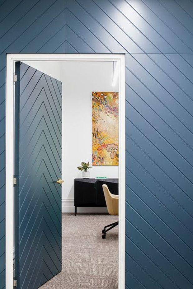 A narrow view through an open interior door into a small office or study. The surrounding walls and door are clad in blue painted diagonal chevron paneling, with white trim around the doorway. Inside is a black desk or credenza, a tan office chair, carpeted flooring, a small plant, and colourful wall artwork.