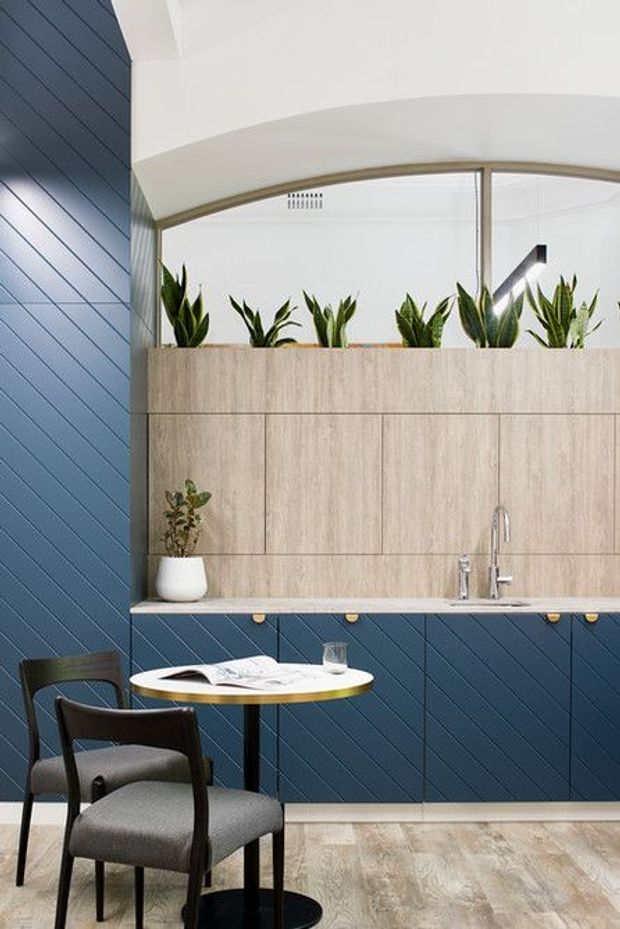 A compact contemporary dining and kitchenette area with a round table and upholstered chairs in the foreground. The space features deep blue diagonal wall panelling and matching lower cabinetry, pale wood-look upper cabinetry, a slim stone or composite benchtop, integrated sink and tapware, a large curved mirror or glazed reflected surface, and planter boxes with green foliage above the joinery.