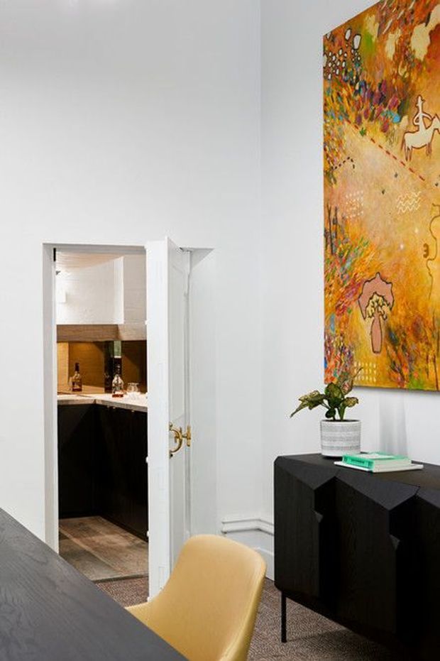 Bright, refined interior view of a dining or living area with white walls, a black geometric sideboard, mustard upholstered chair, dark dining table edge, and a colourful abstract artwork. An open doorway reveals a compact dark-toned kitchen or bar area with timber flooring and a white benchtop.