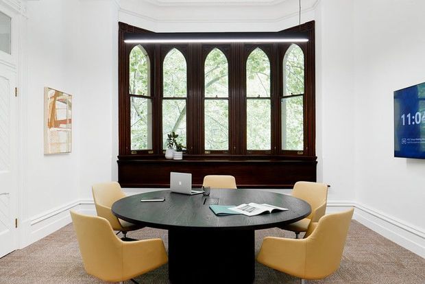 A refined meeting room with a round dark timber conference table, six mustard-yellow upholstered chairs, and a laptop and documents arranged on the table. White walls with decorative moulding surround a prominent dark-stained timber bay window with tall arched panes and a built-in sill. A wall-mounted display is visible on the right, and the room has neutral carpet flooring with soft natural light from the windows.