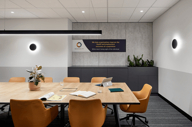 Contemporary commercial meeting room with a large light-wood conference table, warm tan upholstered office chairs, dark built-in storage cabinetry, a textured concrete-look feature wall, linear pendant lighting, and potted greenery. The room has a suspended grid ceiling, carpeted floor, and wall-mounted branding signage.