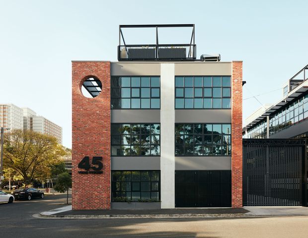 Street-facing exterior of a compact multi-storey commercial or mixed-use building, with red brick end walls, a pale rendered central facade, large dark-framed grid windows, a black garage or entry opening, rooftop terrace railings, and prominent building signage reading 45. The building sits in an urban streetscape with neighbouring apartment and commercial buildings visible around it.