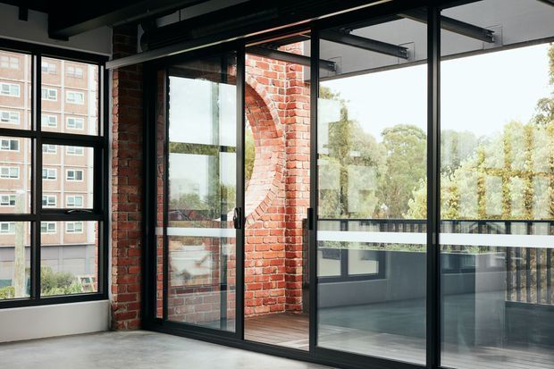 Industrial-style interior or enclosed balcony space with exposed red brick walls, black-framed floor-to-ceiling glazing, a glazed door, exposed dark ceiling structure, and a polished concrete floor. Large windows reveal an adjacent apartment building and trees outside. The space is bright and open, with a strong warehouse-conversion character.