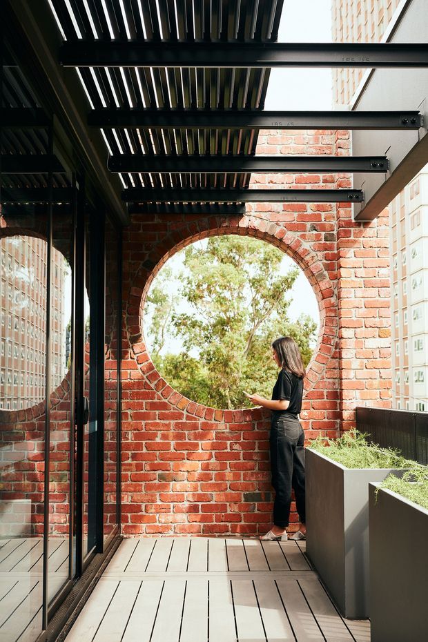 A narrow elevated outdoor terrace or balcony beside a brick building, featuring a circular opening in a red brick wall, black-framed glazing, dark overhead louvres or pergola beams, and built-in planter boxes. The timber-look decking and exposed brick create a warm industrial-heritage character, with trees visible through the opening and neighbouring high-rise buildings in the background.