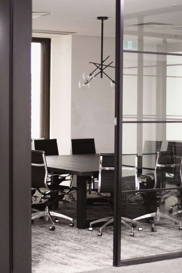 A modern commercial meeting room viewed through dark-framed glass doors and partitions. The room contains a long dark conference table, multiple black office chairs with chrome bases, a contemporary multi-arm pendant light, white walls, a suspended ceiling, and grey patterned carpet. Reflections in the glazing show additional chairs and partition lines.