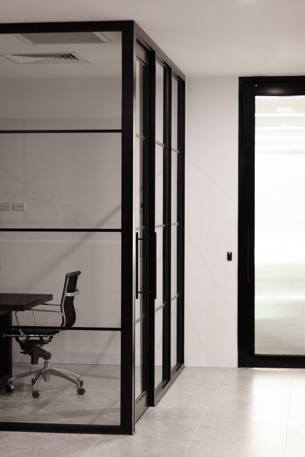 A minimalist commercial office interior with a glazed meeting room enclosed by black-framed glass partitions. A desk and office chair are visible inside the room, while a black-framed frosted-glass door and pale tiled flooring occupy the corridor area.