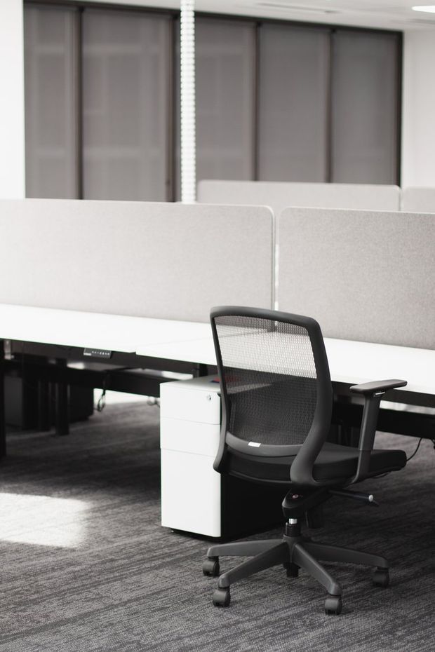 A bright, modern open-plan office workspace with rows of white desks separated by tall light-grey acoustic privacy screens. A black ergonomic mesh office chair sits in the foreground beside a white mobile pedestal, with dark carpet flooring and grey roller blinds along the rear windows.