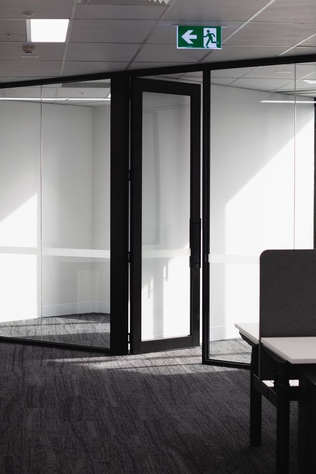Bright modern office interior with full-height glazed partitions, black-framed glass doors, dark patterned carpet, white walls, and a suspended acoustic tile ceiling. A green emergency exit sign is mounted above the doorway, and part of a dark workstation is visible in the foreground.