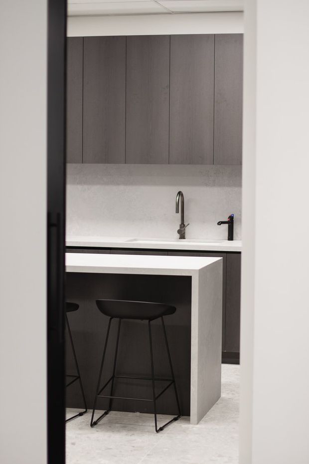 A compact, contemporary kitchen viewed through a doorway, featuring dark vertical wood-grain cabinetry, a light stone or engineered-stone benchtop and island, integrated sink with a curved metal tap, black faucet, and two black minimalist bar stools. The room has a restrained monochromatic palette and a clean, functional composition.