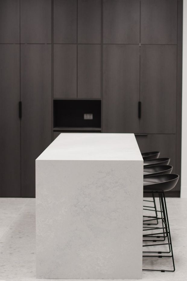 A minimalist contemporary kitchen featuring a large white stone island with waterfall sides and seating for several people. The island is positioned in front of full-height dark charcoal cabinetry with integrated handles and a recessed black appliance niche. Black bar stools line one side, and the pale floor and restrained palette create a refined, monochromatic look.