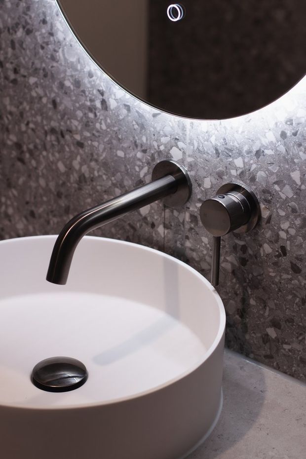Close-up of a refined bathroom vanity featuring a white round vessel basin, wall-mounted dark metal tapware, a large circular mirror, and a grey terrazzo-style wall surface. The composition is modern and minimalist, with a soft neutral palette and sculptural curved forms.